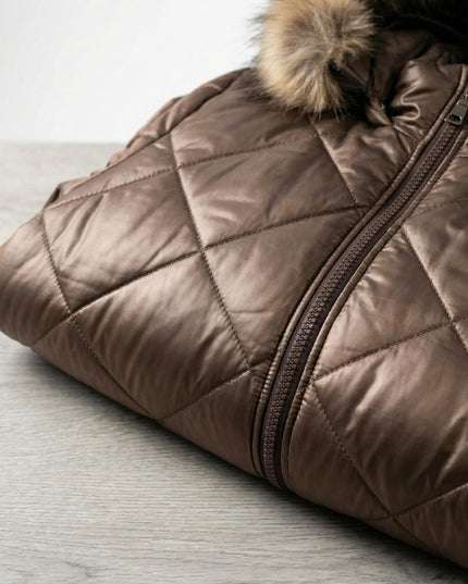 Сгънато кафяво зимно яке с диамантено капитониране върху дървена повърхност, показващо закопчаването с цип и част от качулката с естествен косъм.