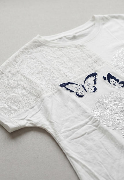 Детайл отблизо на бяла лятна памучна туника, показващ фин дантелен панел и изящна апликация с тъмносиня пеперуда и сребристи цветя.