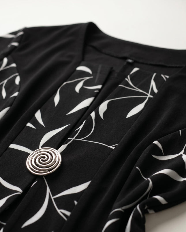 Детайл отблизо на черна официална рокля големи размери, показващ ефектен бял листен мотив и блестяща сребриста спираловидна брошка.