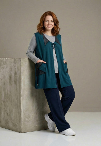 Дълъг разкроен макси елек в масленозелено с еко кожа и връзки, заснет в цял ръст върху модел, позираща до сив бетонен куб.