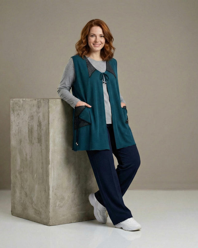 Дълъг разкроен макси елек в масленозелено с еко кожа и връзки, заснет в цял ръст върху модел, позираща до сив бетонен куб.