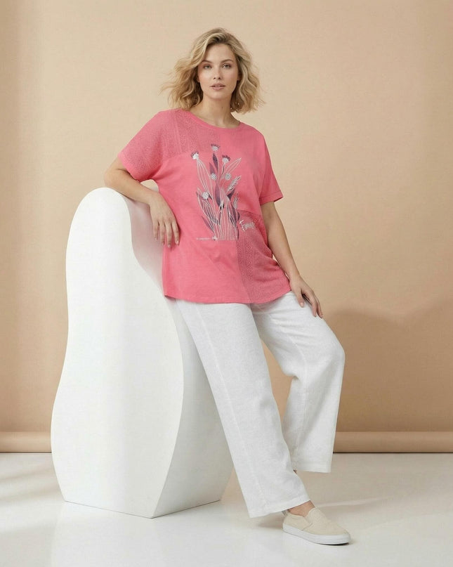 Дамска лятна туника в коралов цвят с флорална щампа и дантела, представена в цял ръст от модел, съчетана с бели панталони на бежов фон.