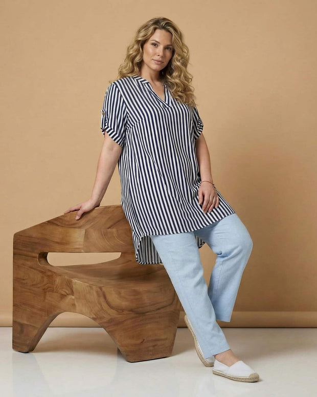 Жена облечена с раирана туника в голям размер със свободна кройка комбинирана със светъл панталон в студио с топъл бежов фон и дървен декоративен елемент