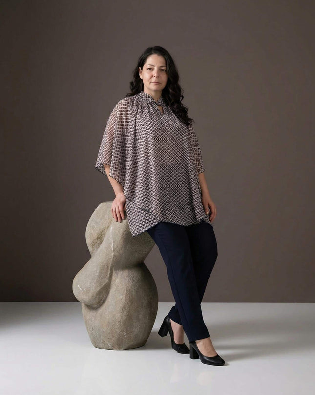 Ефирна шифонена туника с геометричен принт и пончо ефект, представена в цял ръст върху модел до каменна скулптура.