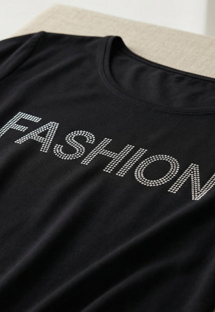 Детайл отблизо на черна асиметрична туника, показващ ефектен декоративен надпис FASHION, изработен от блестящи камъчета.
