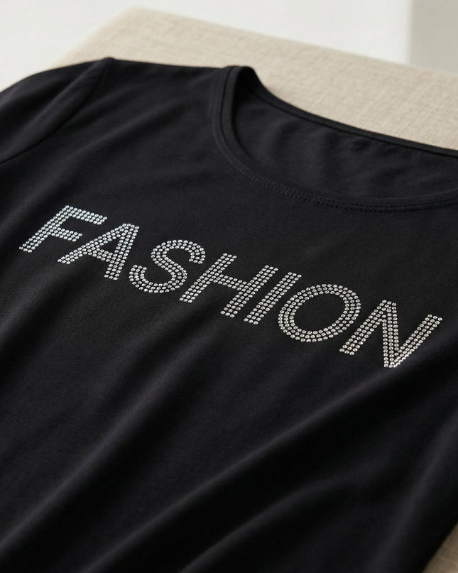 Детайл отблизо на черна асиметрична туника, показващ ефектен декоративен надпис FASHION, изработен от блестящи камъчета.