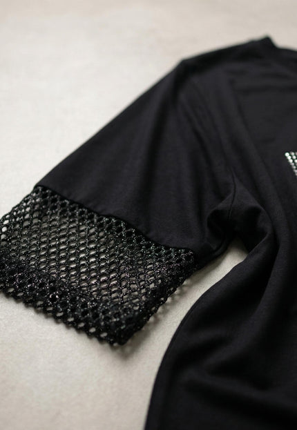 Детайл отблизо на елегантна черна асиметрична туника, показващ ръкава с блестяща мрежа и част от надписа с камъчета.
