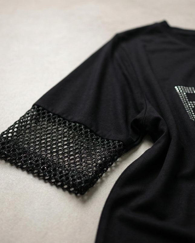 Детайл отблизо на елегантна черна асиметрична туника, показващ ръкава с блестяща мрежа и част от надписа с камъчета.