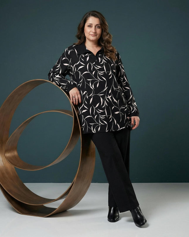 Елегантна макси туника с абстрактен принт и черна яка, представена в цял ръст. Моделът е облечен с черен панталон и позира до декоративен елемент.