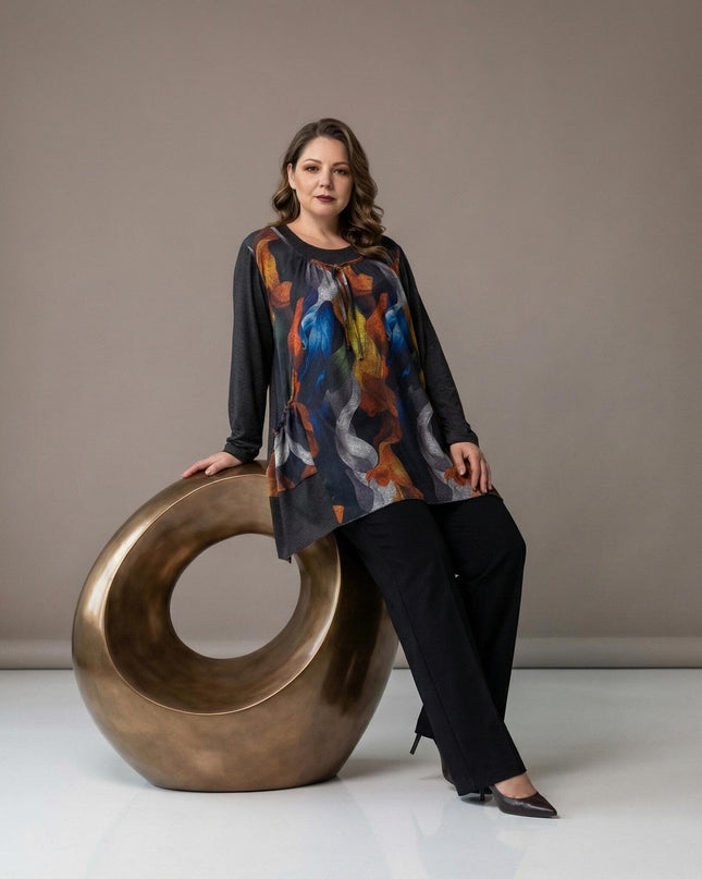 Елегантна разкроена туника с абстрактен цветен принт и асиметричен подгъв, заснета в цял ръст върху модел в седяща поза.