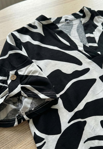 Детайл отблизо на лятна макси туника с черно-бял принт. Фокус върху навития къс ръкав, фиксиран с дървено копче, заснет като плосък кадър върху маса.