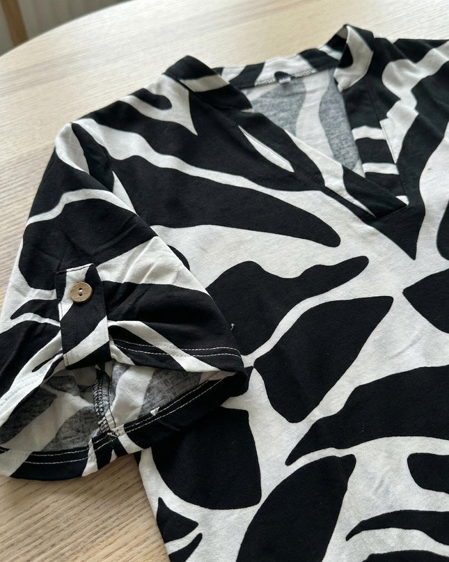 Детайл отблизо на лятна макси туника с черно-бял принт. Фокус върху навития къс ръкав, фиксиран с дървено копче, заснет като плосък кадър върху маса.