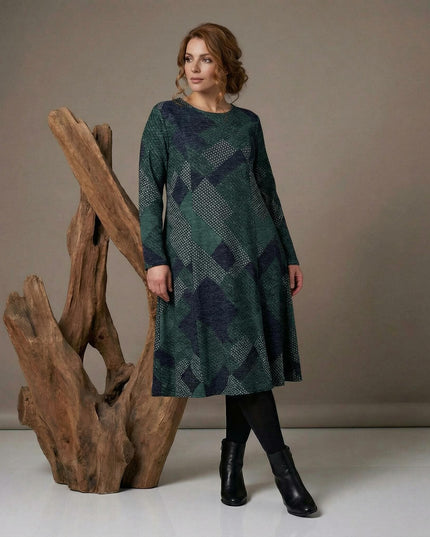 Масленозелена разкроена рокля с геометричен пачуърк и дълъг ръкав, представена в цял ръст до декоративен дървен елемент.