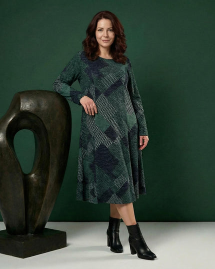 Масленозелена разкроена рокля с геометричен пачуърк и дълъг ръкав, представена в цял ръст върху модел до абстрактна скулптура.