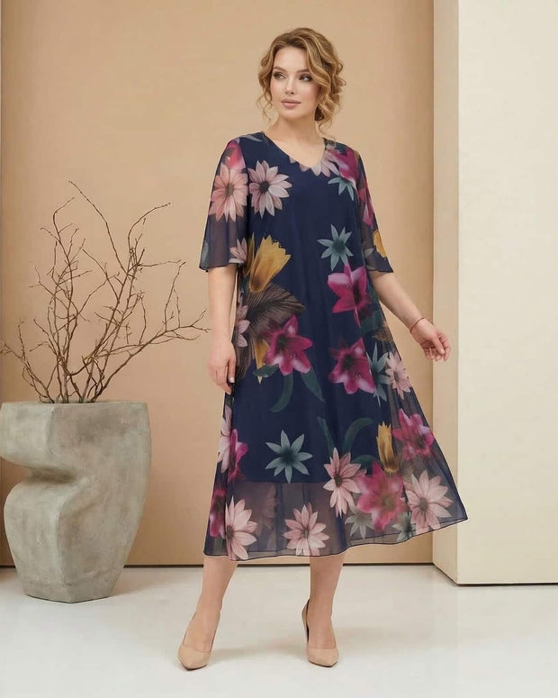 Жена облечена с официална шифонена рокля в голям размер с флорален принт в елегантна студийна обстановка със светъл фон и декоративна ваза с клонки