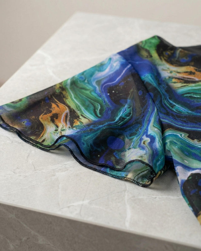 Детайл отблизо на официална шифонена рокля големи размери, показващ къс ръкав, преливащи шарени тонове и зашити ръбове, разположена върху светъл мраморен фон.