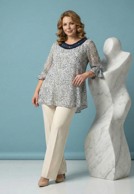 Стилна бяла макси туника с флорален принт и деколте с перли, представена от модел в цял ръст, опиращ се на бяла скулптура.