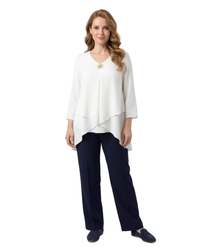 Официална бяла макси туника с асиметрична кройка на пластове и брошка, показана в цял ръст на бял фон, съчетана с тъмносин панталон.