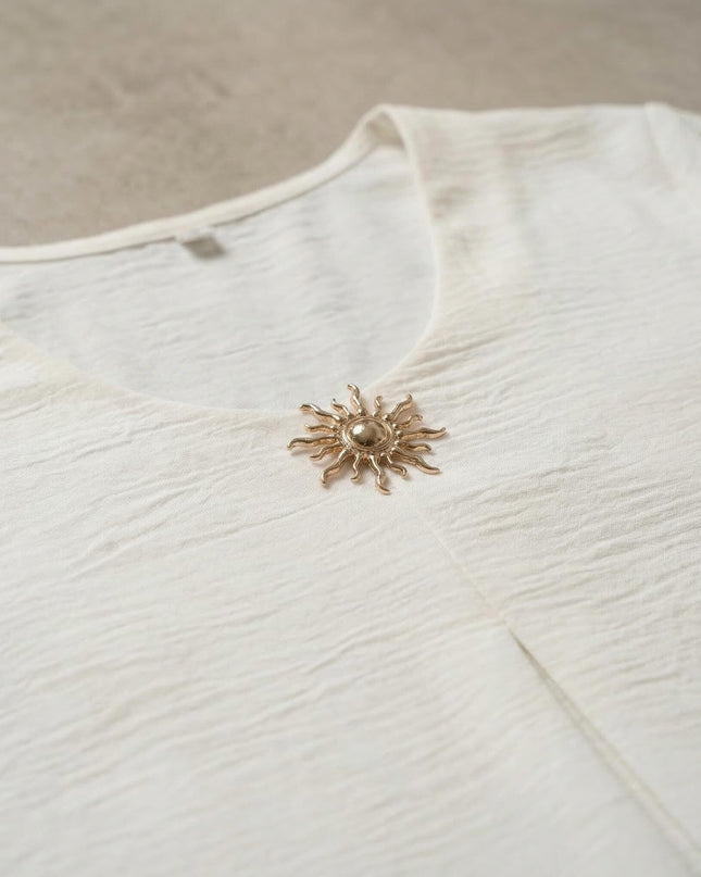 Детайл отблизо на официална бяла макси туника с релефна материя и ефектна златна брошка във формата на слънце на деколтето.