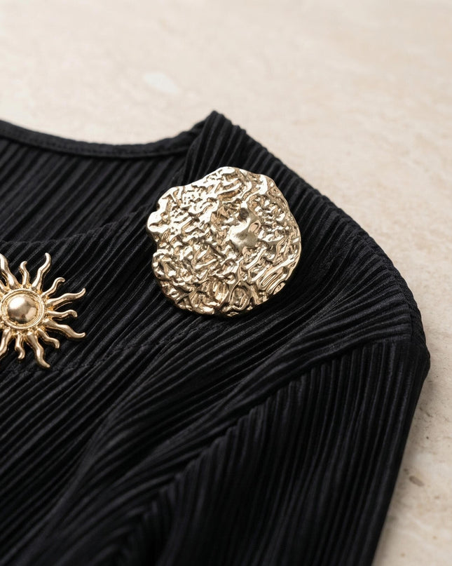Детайл отблизо на черна макси туника, акцентиращ върху фино плисираната материя солей и елегантните златисти брошки.