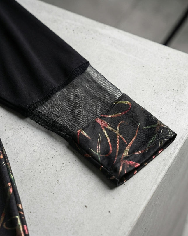 Детайл от ръкава на официална черна туника, показващ мрежест елемент и маншет с цветен абстрактен принт върху сива повърхност.