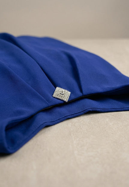 Детайлен кадър на официална туника в кралскосин цвят, акцентиращ върху блестящата квадратна брошка и елегантните набори на шифонената материя.