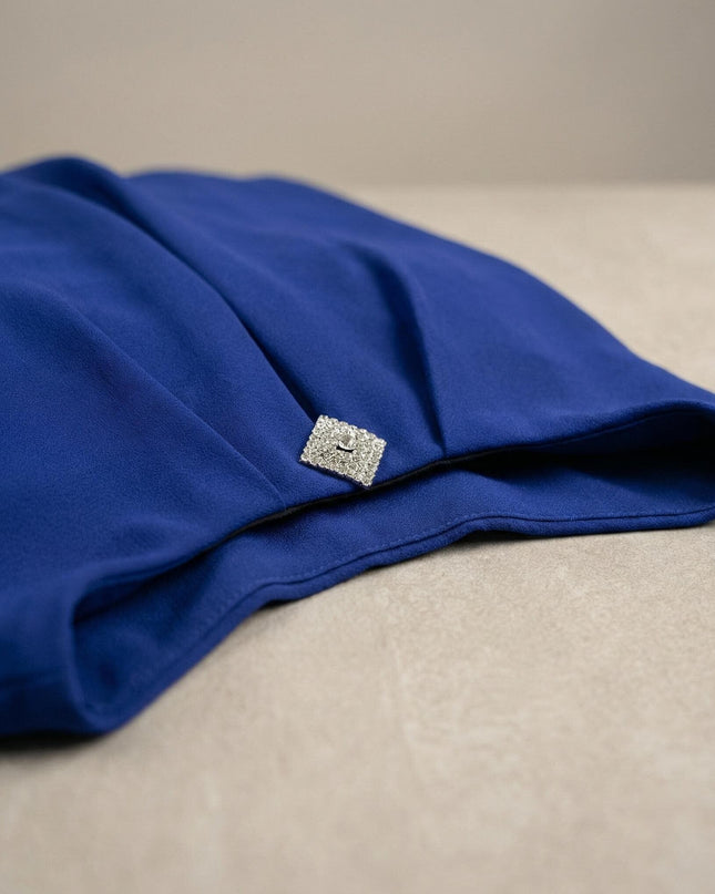 Детайлен кадър на официална туника в кралскосин цвят, акцентиращ върху блестящата квадратна брошка и елегантните набори на шифонената материя.
