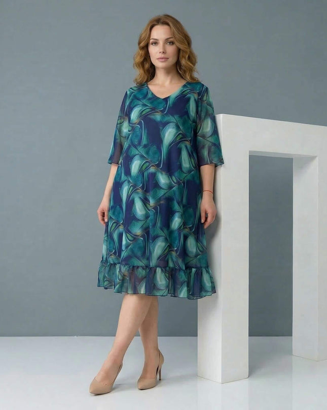 Официална шифонена рокля големи размери в синьо-зелени преливащи тонове, представена в цял ръст с V-образно деколте и волан на подгъва.