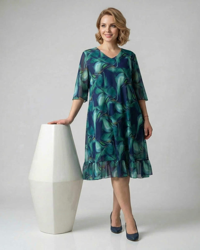 Официална шифонена рокля големи размери в преливащи сини и зелени тонове, цял ръст на бял фон, с V-образно деколте и волан.