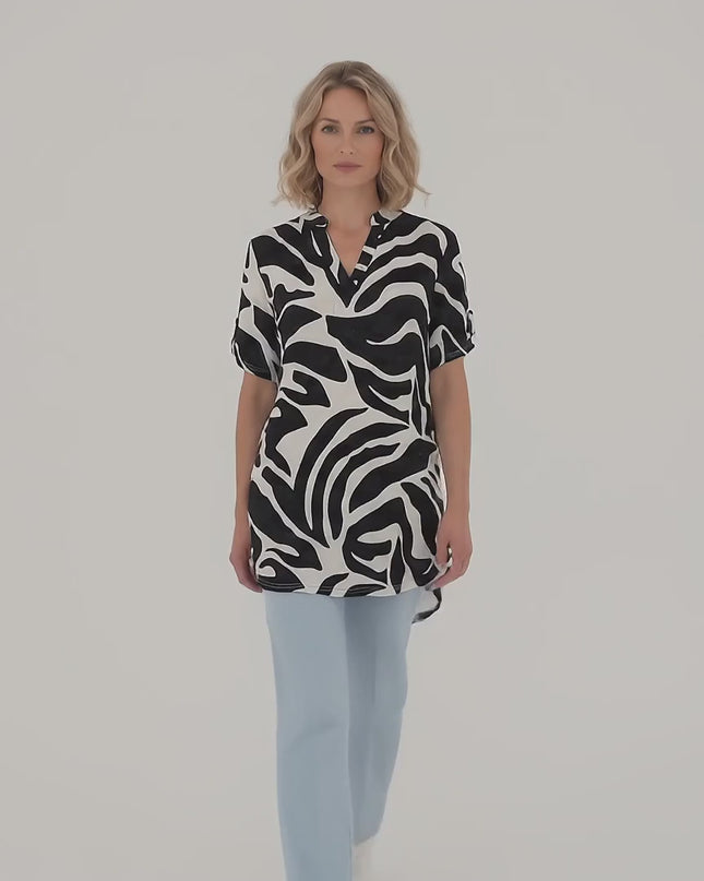 Плюс размер модел носи Макси лятна туника в черно и бяло с асиметрична кройка и върви бавно към камерата на неутрален фон в студийна среда