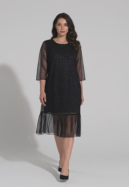 Видео в студийна среда, показващо плюс размер модел на неутрален фон, който върви бавно към камерата и е облечен в Официална разкроена макси рокля в черно от шифон с камъни и 7/8 ръкави