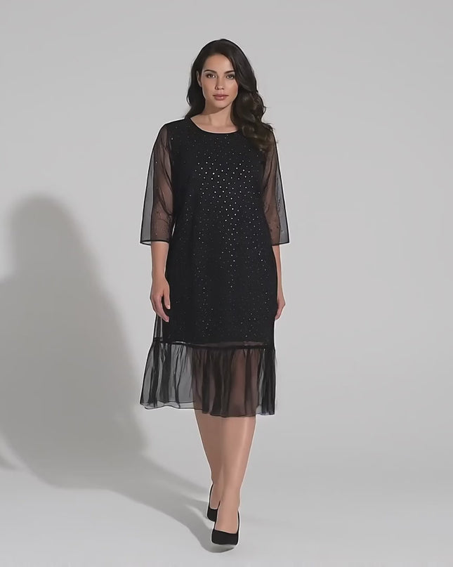 Видео в студийна среда, показващо плюс размер модел на неутрален фон, който върви бавно към камерата и е облечен в Официална разкроена макси рокля в черно от шифон с камъни и 7/8 ръкави