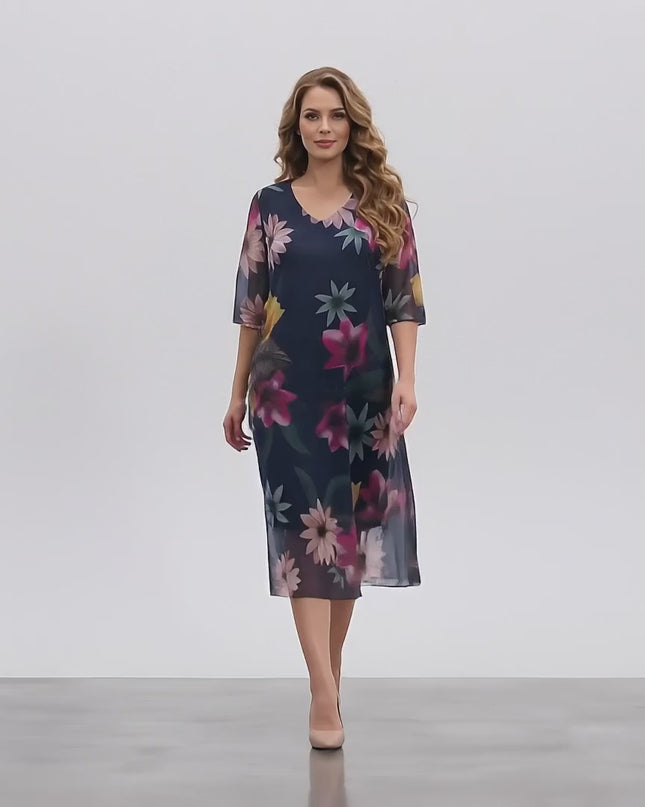 Макси модел се разхожда бавно към камерата в студийна среда с неутрален фон, носейки Официална тъмносиня макси рокля с ефирен флорален шифон