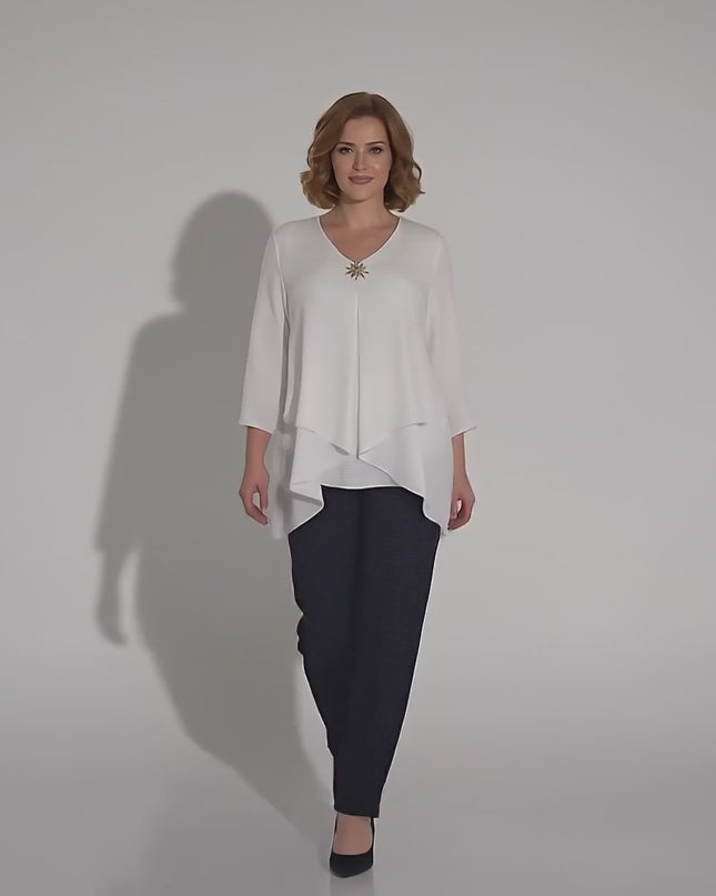 Модел плюс размер, облечен с Официална бяла макси туника с асиметрична кройка и брошка, който върви бавно към камерата в студийна среда на неутрален фон