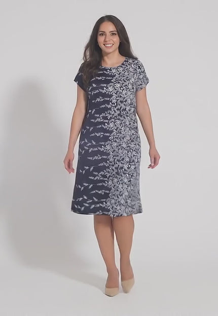 Плюс сайз модел, облечена в Лятна тъмносиня рокля с флорални мотиви и права кройка, се разхожда бавно към камерата в студио с неутрален фон