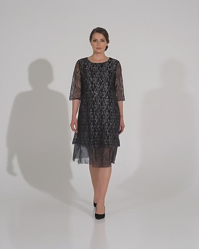 Плюс сайз модел върви бавно към камерата в студийна среда на неутрален фон, облечена в Официална черна макси рокля с ефектна дантела