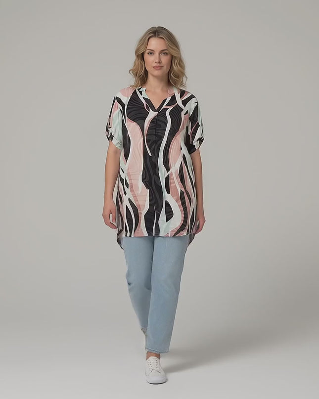 Плюс размер модел се разхожда бавно към камерата в студийна среда, облечена в Дълга лятна макси туника с шарен принт и асиметрична кройка