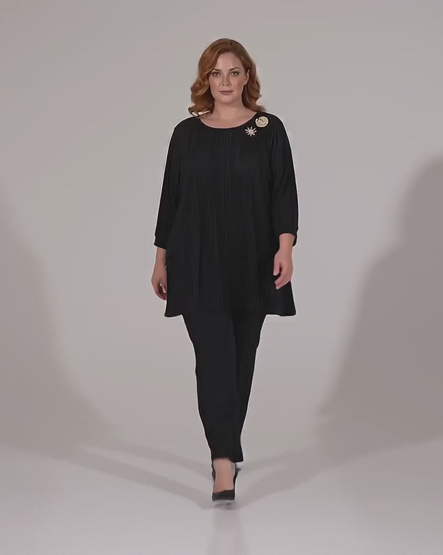 Видео в студийна среда, на което макси модел върви бавно към камерата, облечена в Официална черна макси туника от солей с брошка