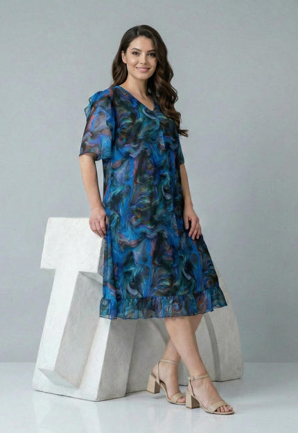 Шарена официална рокля големи размери с абстрактен принт и шифонени елементи, показана в цял ръст върху модел до бял декор.