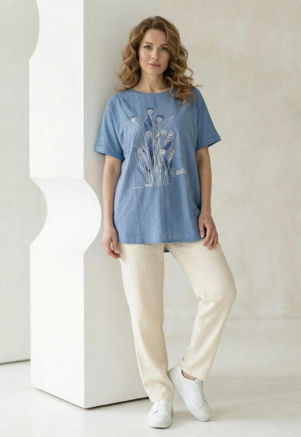 Синя лятна туника големи размери с флорални мотиви и дантела, съчетана със светъл панталон. Снимка в цял ръст на модел, позиращ в студио.