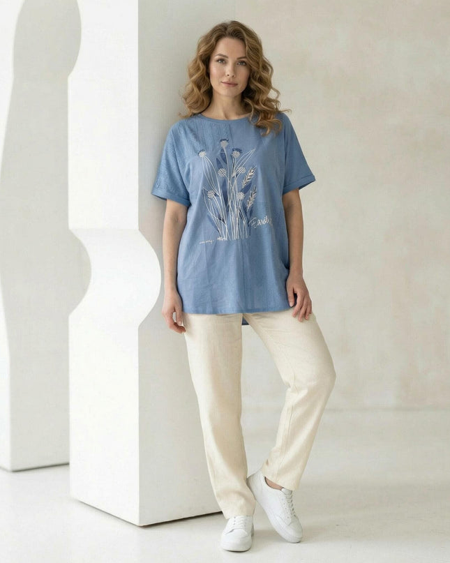 Синя лятна туника големи размери с флорални мотиви и дантела, съчетана със светъл панталон. Снимка в цял ръст на модел, позиращ в студио.