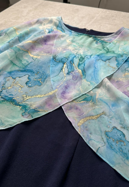 Детайл отблизо на горната част на тъмносиня официална рокля, акцентиращ върху ефирното шифонено наметало с красив абстрактен принт.